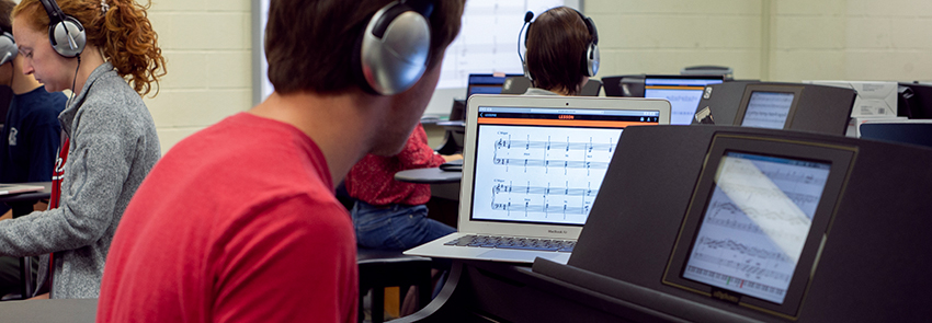 A student wearing headphones consults sheet music displayed on a laptop and a tablet.