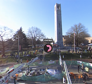 Runners begin the Krispy Kreme Challenge in front of the NC State Bell Tower on a sunny day. There is a donut icon in front of the image from the VR experience.