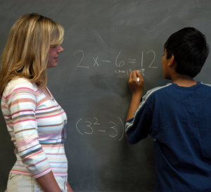 Teacher helps student work on math problem on a chalkboard.