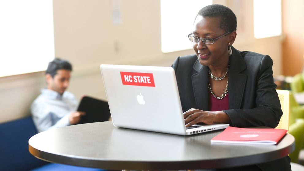 Woman sits at a table with a laptop and notebook. Laptop has NC State sticker.
