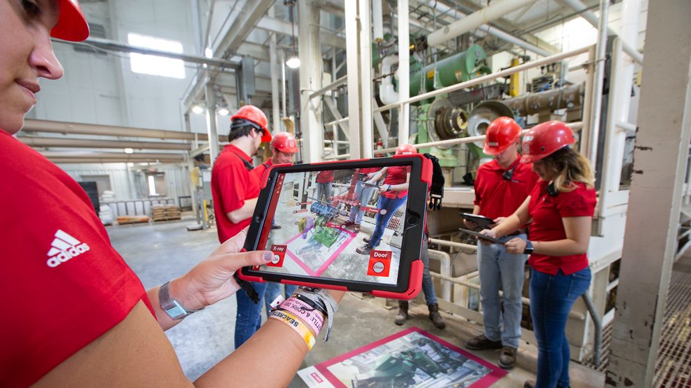 Taken inside the feed mill, a person holds a tablet with the AR experience loaded. There are other students gathered around a map of the Feed Mill and on the screen you can see the AR experience on the mat.