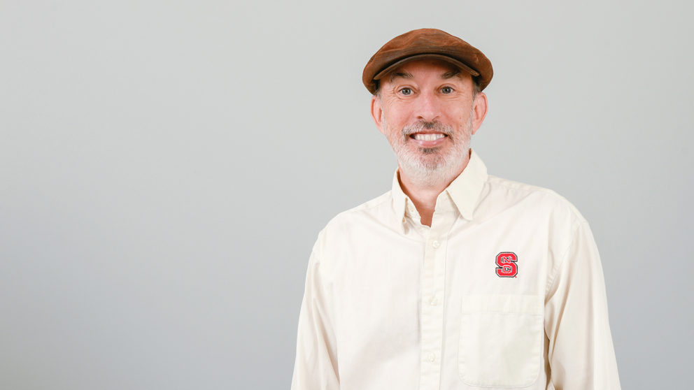 Portrait of David Howard against a white background. He is pictured wearing an NC State shirt and a brown hat.