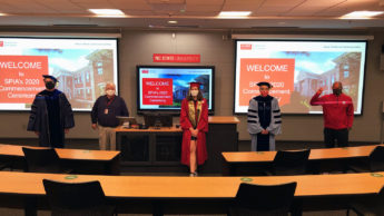 Five people stand physically distanced in a classroom. Projectors and a television display the SPIA 2020 Commencement Ceremony