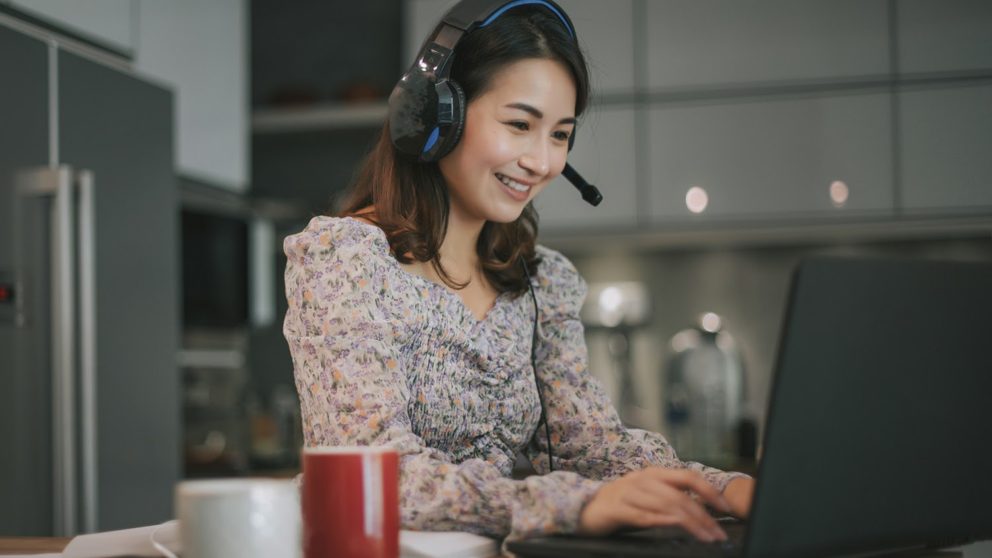 Woman smiles while working on computer and wearing a headset.