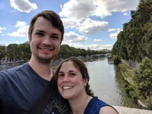 Two people smile at a camera overlooking a river in Rome.