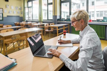 Happy mature female teacher writing notes in her notebook while using a computer in the classroom.