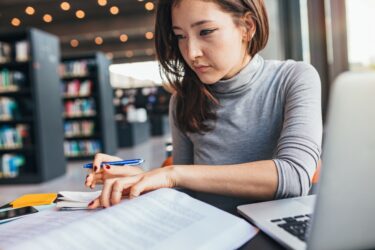 Young woman studying on a book and taking down note while sitting at the library desk. Asian female student preparing for final exams.